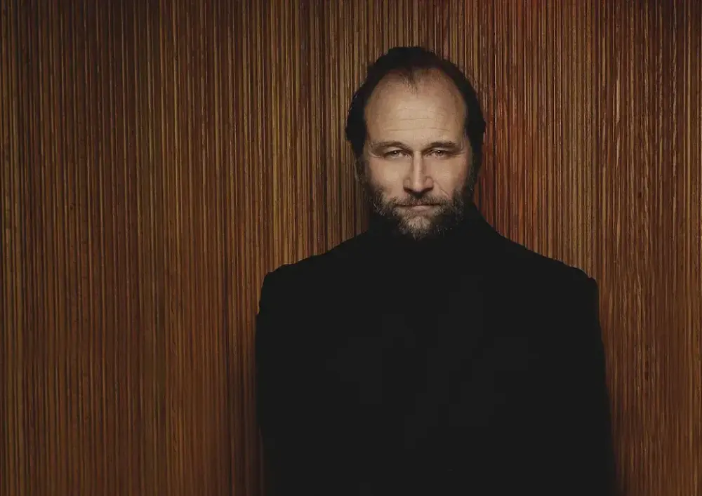 Stein Torleif Bjella i mørke kle står framom ein trevegg