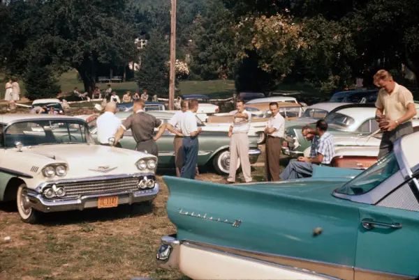 Samling av mennesker rundt amerikanske veteranbiler parkert på gress.