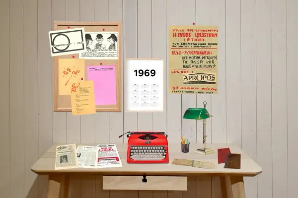 ein skrivepult med skrivemaskin og aviser og bøker. På veggen heng det ein plakat og ei oppslagstavle med flygeblad.