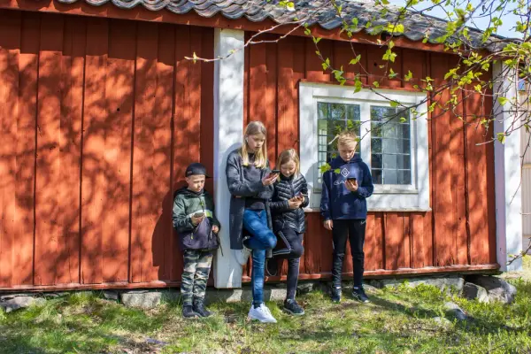Fyra barn står tillsammans vid ett rött hus och tittar på sina mobiler.