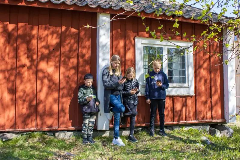 Fyra barn står tillsammans vid ett rött hus och tittar på sina mobiler.