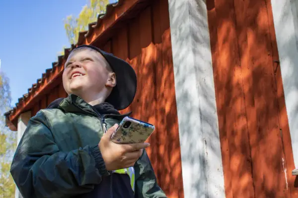 En pojke i keps står vid ett rött hus. Han håller i sin telefon och tittar leendes upp åt sidan.