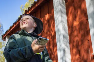 En pojke i keps står vid ett rött hus. Han håller i sin telefon och tittar leendes upp åt sidan.