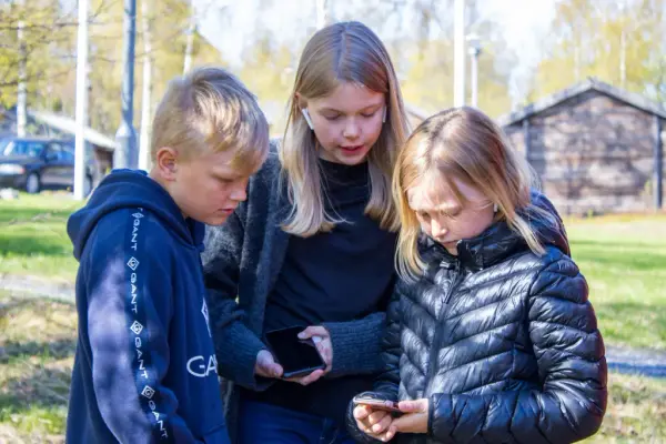 Tre barn står tillsammans utomhus och tittar ner på en telefon.