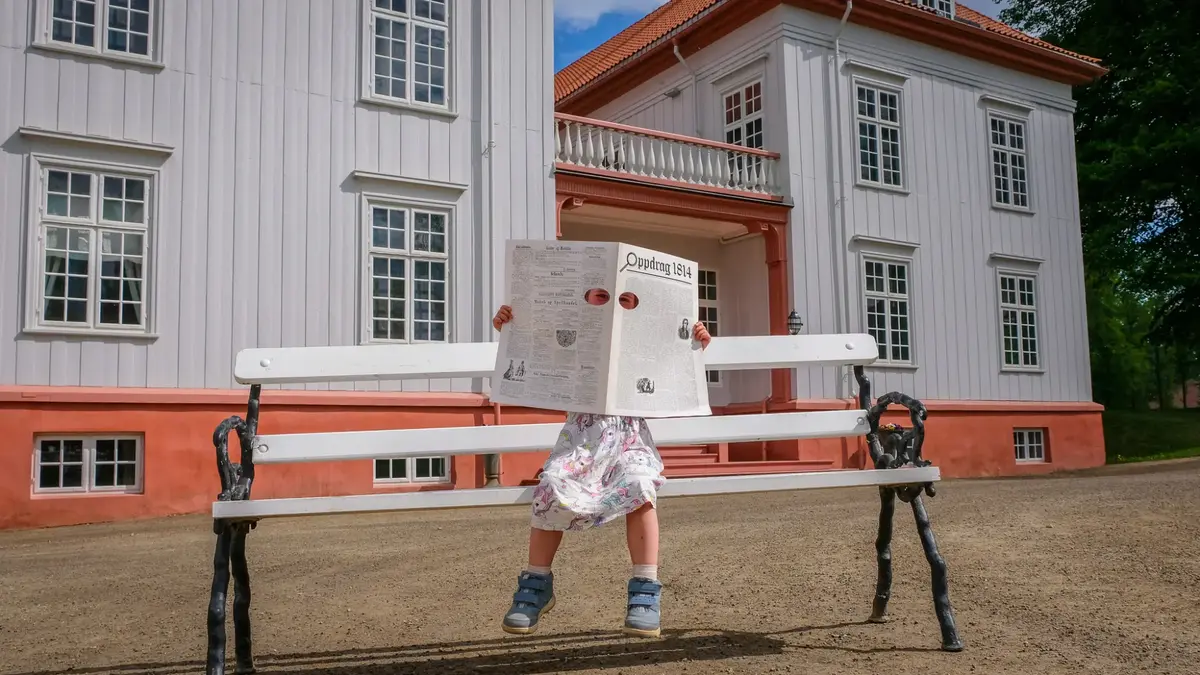 Barn på benk foran Eidsvollsbygningen leser et stort ark med kikkehull, del av aktiviteten «Hemmelig budbringer».