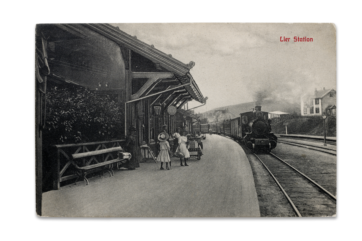 Lier stasjon ca. 1910. Toget tilhører Drammenbanen og er på vei i retning Kristiania..Den flotte stasjonsbygningen i sveitserstil fra 1870, er tegnet av jernbanens arkitekt Georg Andreas Bull. Det er dessuten det første kjente arkitekttegnede huset i Lier.