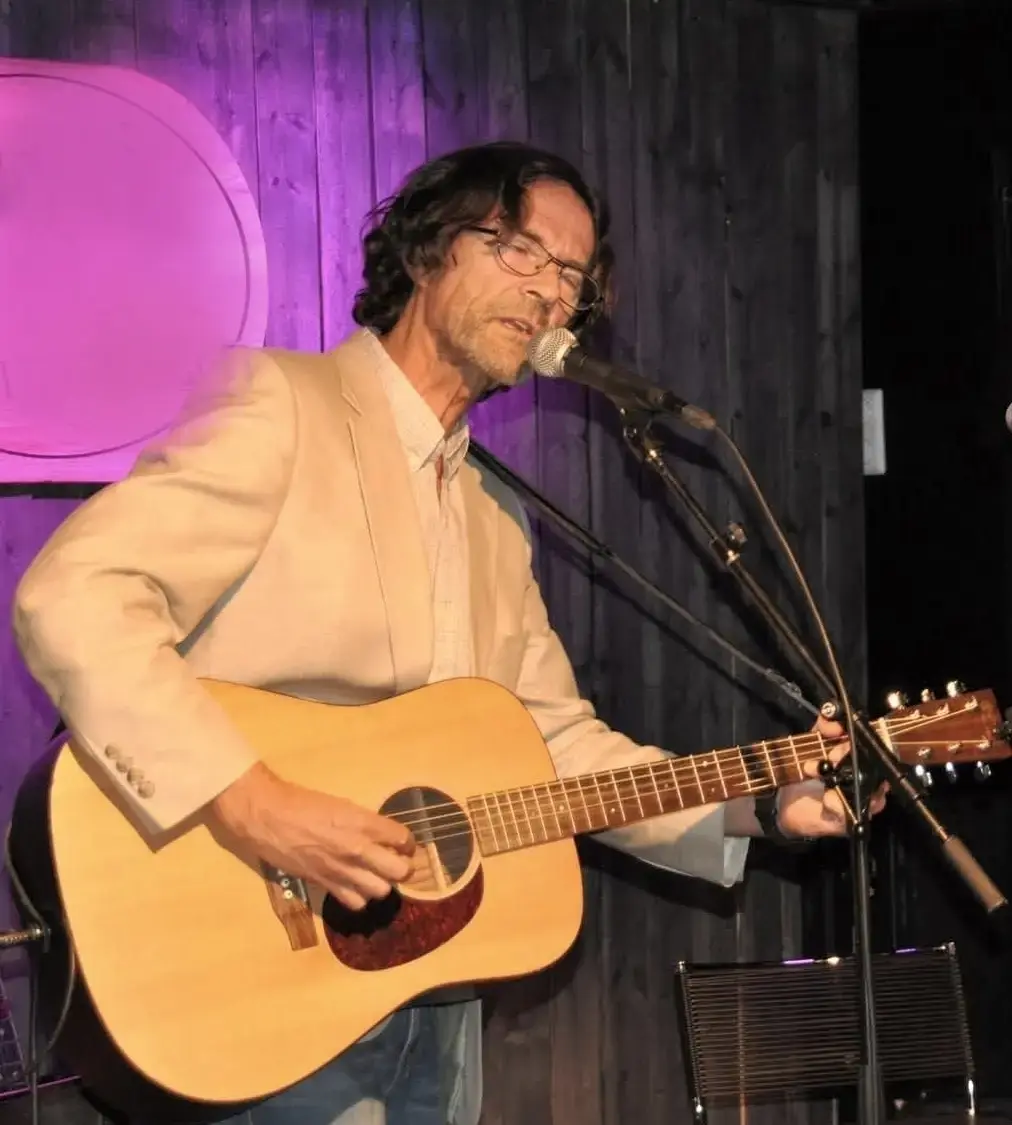 Roar Vea opptrer med akustisk gitar og mikrofon på en innendørs scene, iført beige blazer og briller.