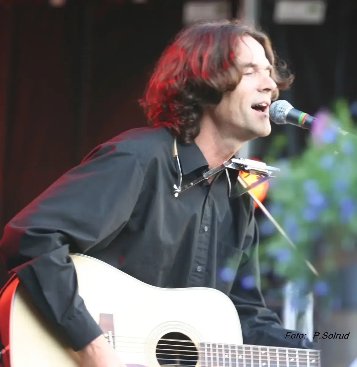 Roar Vea synger og spiller gitar under en livekonsert, med munnspillstativ og mikrofon foran seg.