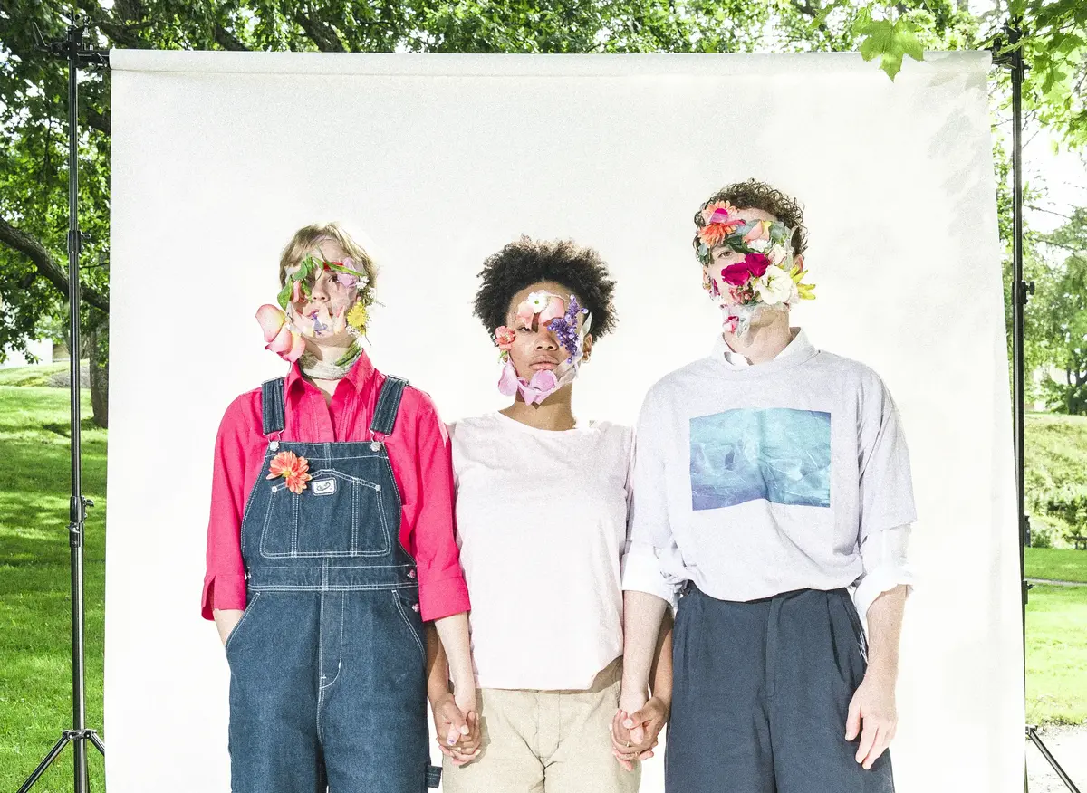 Tre personer på rad håller varandra i hand, de har blommor i ansiktet