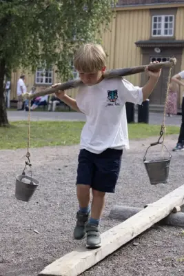 Gutt balanserer på planke med melkespann over skuldrene på Norsk Folkemuseum.