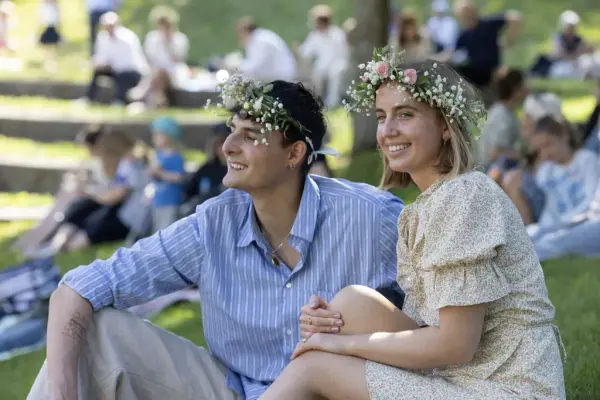 To unge voksne med blomsterkranser sitter i gresset og nyter Sankthansfestivalen på Norsk Folkemuseum.