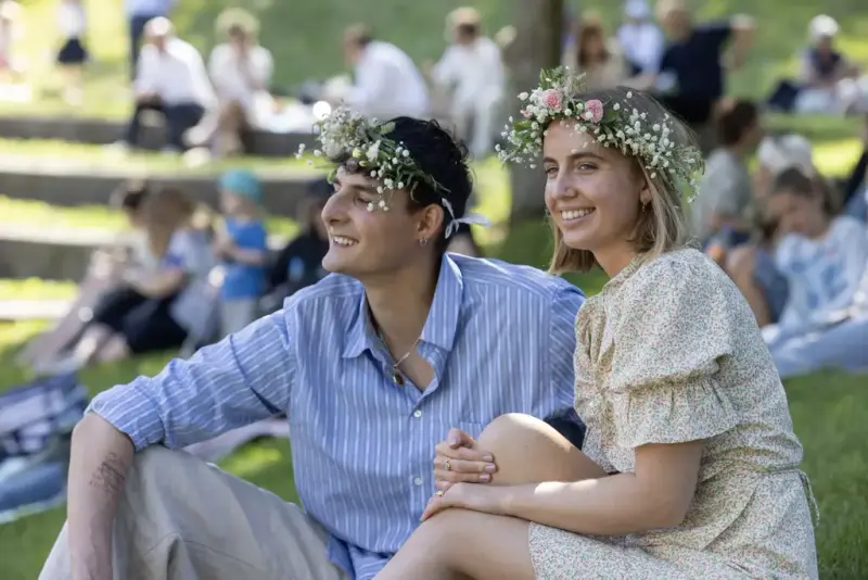 To unge voksne med blomsterkranser sitter i gresset og nyter Sankthansfestivalen på Norsk Folkemuseum.