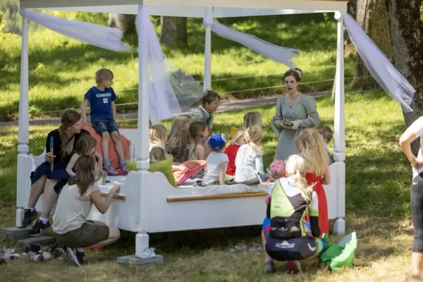 Barn og foreldre sitter i en kjempestor himmelseng ute i parken. En kvinne i gammeldags kjole forteller en historie. 