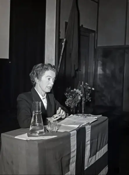 Svart-hvitt bilde av Rakel Sewerin i en blazer og hvit skjorte, bak ett skrivebord med dokumenter, blomster og det norske flagget hengende foran.