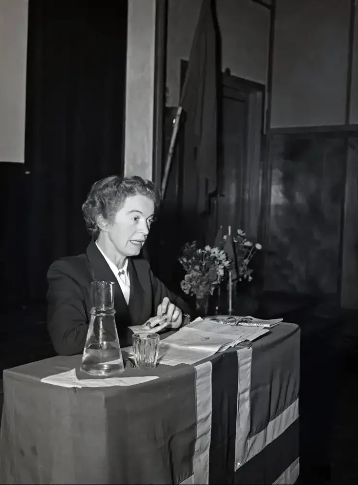 Svart-hvitt bilde av Rakel Sewerin i en blazer og hvit skjorte, bak ett skrivebord med dokumenter, blomster og det norske flagget hengende foran.