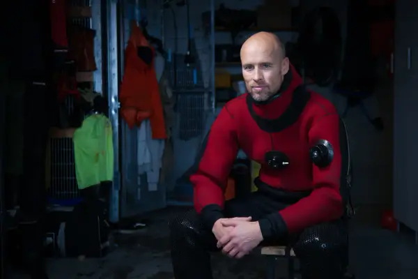 Portrett av Even Moland i en rød dykkerdrakt, sittende i en mørk garderobe.