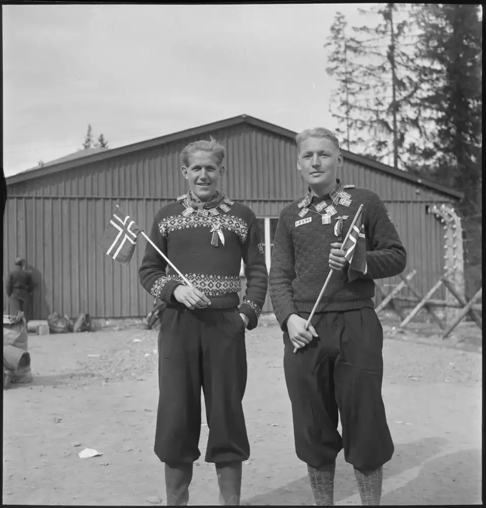 Svart/hvitt fotografi av to mannlige fanger som ser rett i kamera. Begge har norske flagg i hendene og har mange, mindre norske flagg i tråd rundt halsen. De smiler forsiktig.