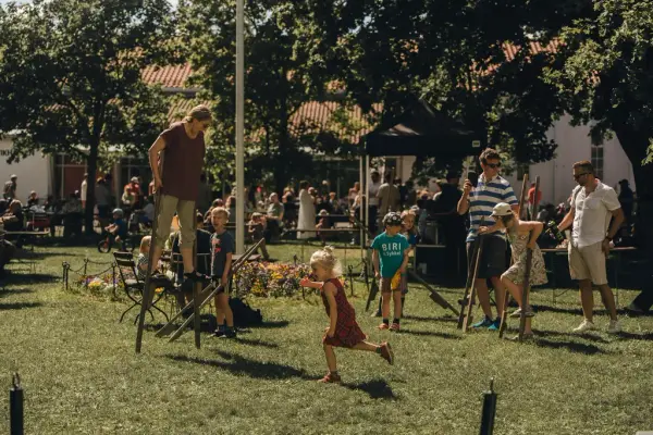 Barn og voksne går på stylter sammen på en sommerlig grønn plen, med en flaggstang og et blomsterbed i bakgrunnen. Mange folk i bakgrunnen av bildet. 