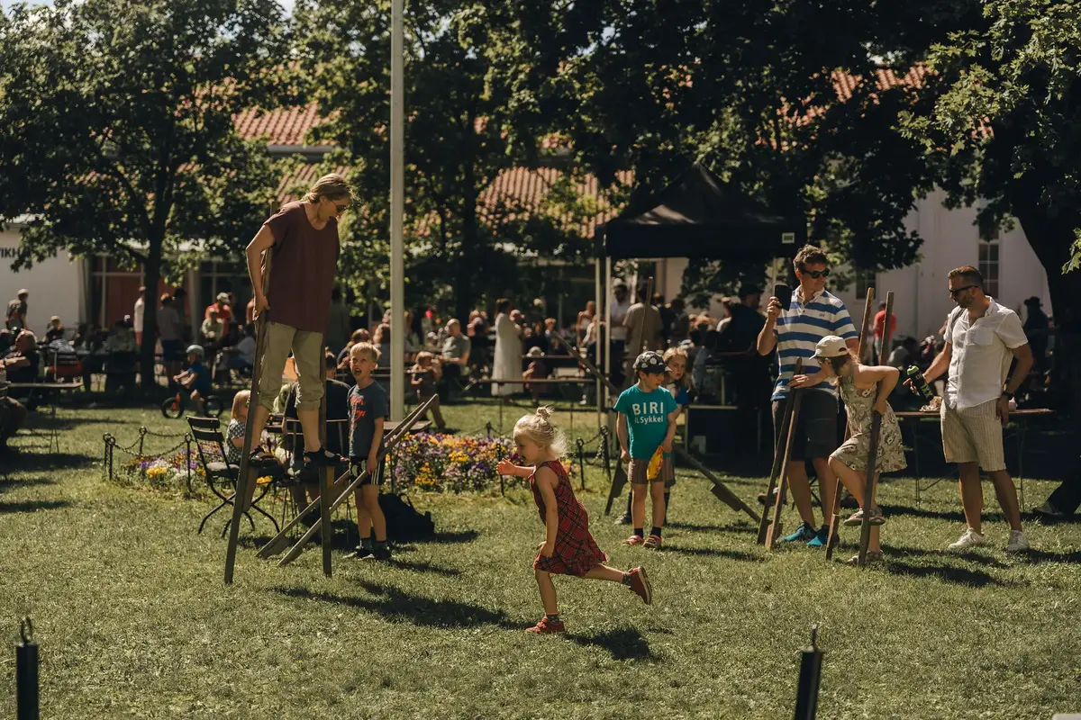 Barn og voksne går på stylter sammen på en sommerlig grønn plen, med en flaggstang og et blomsterbed i bakgrunnen. Mange folk i bakgrunnen av bildet. 