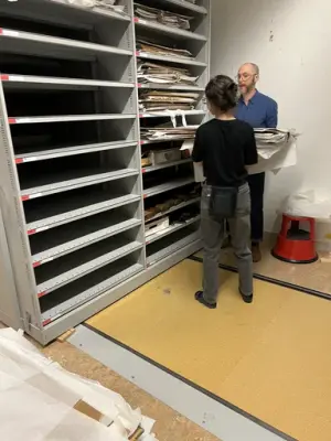 Två personer lyfter skolplanscher in i en magasinshylla