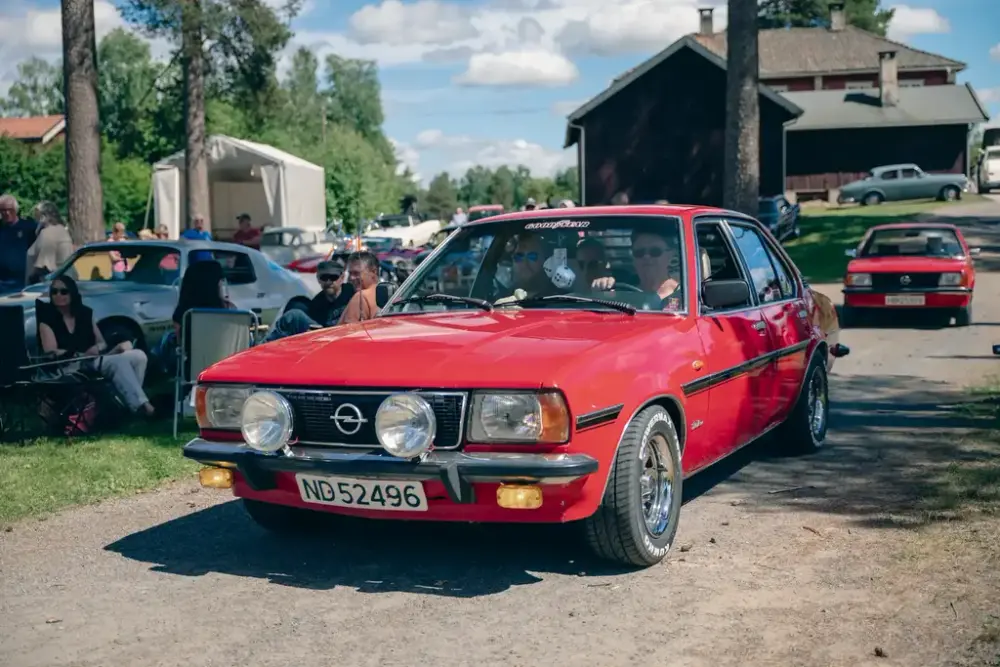 Rød Opel med rallylykter kjører gjennom området under Veterandag på Glomdalsmuseet, omgitt av publikum og klassiske biler