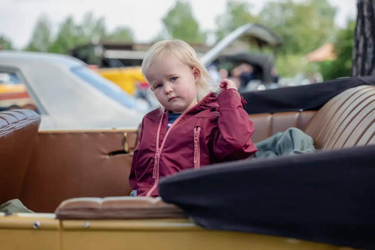 Barn med rød jakke sitter i baksetet på en gammel bil og ser nysgjerrig mot kamera.