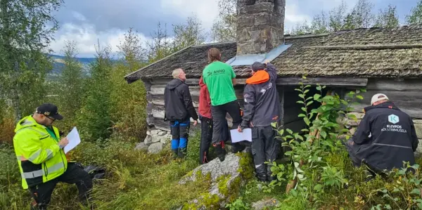 Seks personer står utenfor et gammelt tømmerhus