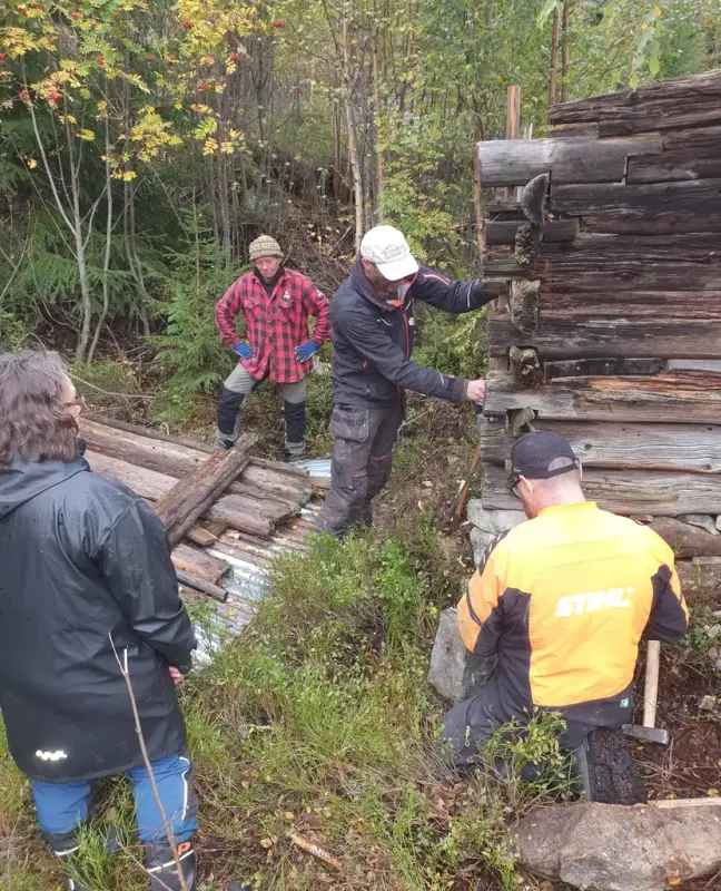 Fire personer står ute i skogen ved et gammelt tømmerbygg