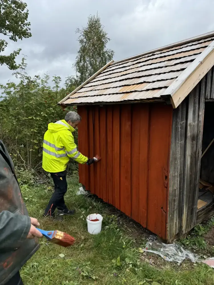 En mann iført en gul refleksjakke maler et skur rødt