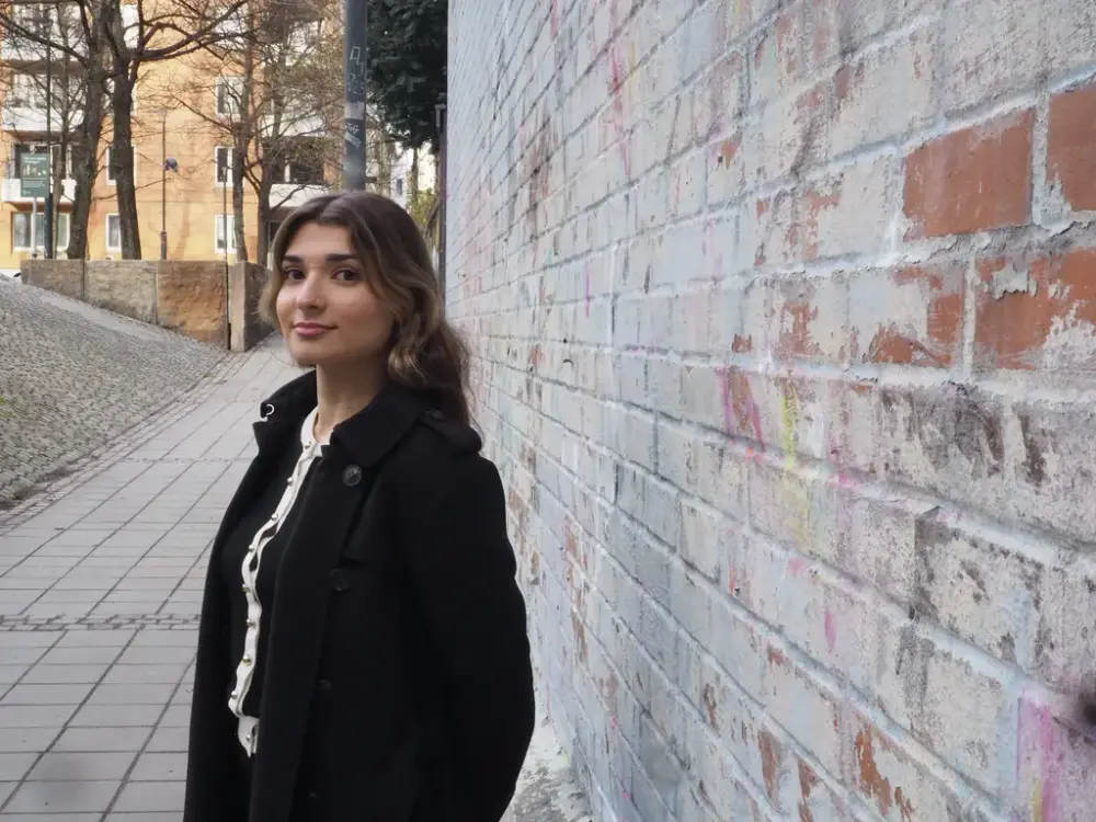 Portrett av en kvinne med sort jakke som står inntil en murvegg på høyre side. Boligblokk i bakgrunn.