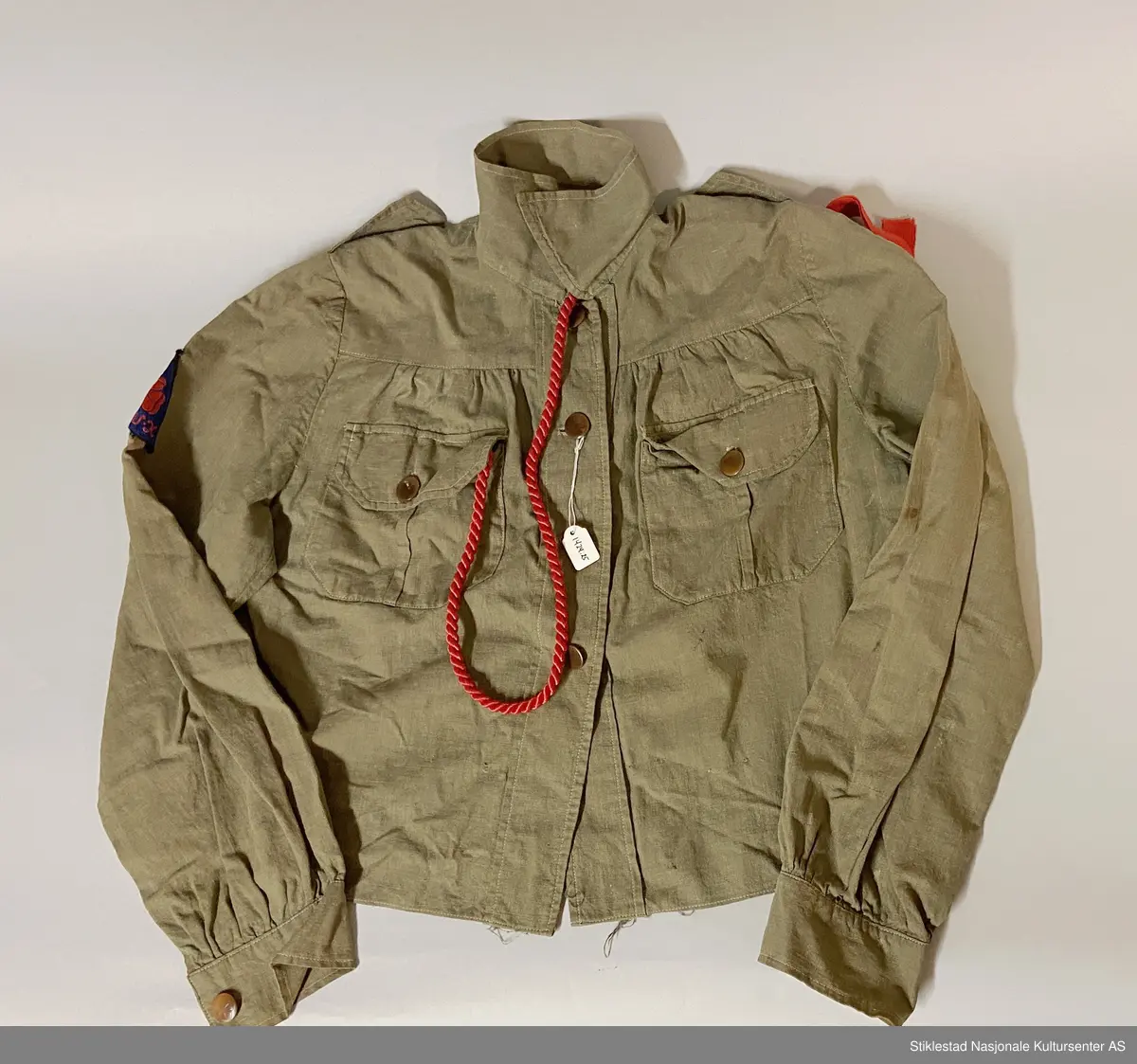 Speideruniform fra 1960-tallet. Dressen er i bomull og består av jakke, skjørt og lue (båtlue) i militærgrønn farge. Jakken har et påsydd stoffmerke på høyre arm: en blå trekant med trekløver og teksten KFUK - ("Kristelig Forening for Unge Kvinner"). På venstre skulderklaff er det festet et rødt bånd (dobbelt) med teksten «Verdal 1» (troppsgrad?). Rød snor i front for fløyte (denne mangler). Skjørtet er kort og "snurpet" sammen i midjen med strikk, to lommer foran. Merket K.K. på innsiden av ene lomme. Lue med påmontert merke i metall; trekløver i trekant. Innskripsjoner på innsiden av luen. Lue, militærgrønn, men annen grønn nyanse enn skjørt og jakke.