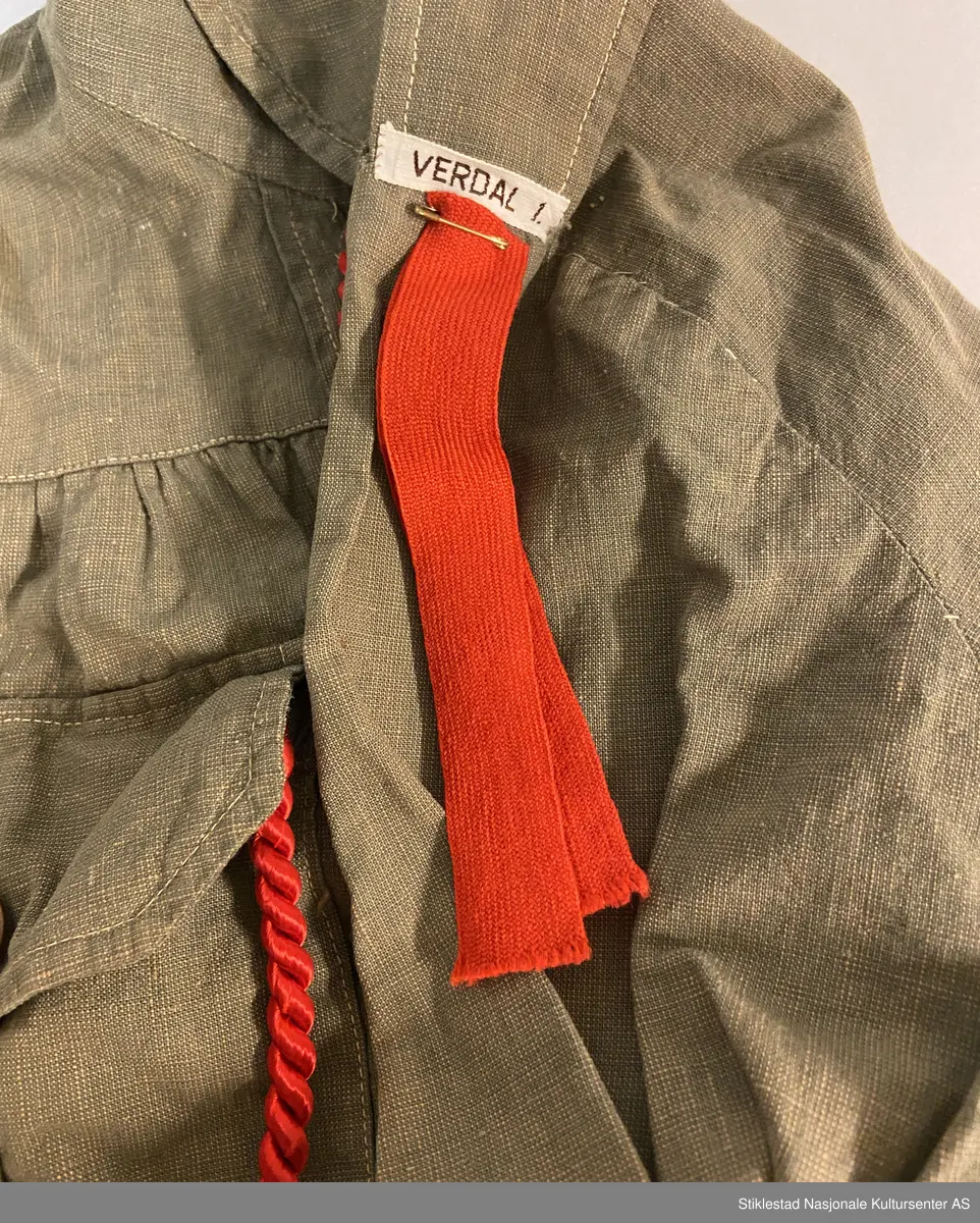 Speideruniform fra 1960-tallet. Dressen er i bomull og består av jakke, skjørt og lue (båtlue) i militærgrønn farge. Jakken har et påsydd stoffmerke på høyre arm: en blå trekant med trekløver og teksten KFUK - ("Kristelig Forening for Unge Kvinner"). På venstre skulderklaff er det festet et rødt bånd (dobbelt) med teksten «Verdal 1» (troppsgrad?). Rød snor i front for fløyte (denne mangler). Skjørtet er kort og "snurpet" sammen i midjen med strikk, to lommer foran. Merket K.K. på innsiden av ene lomme. Lue med påmontert merke i metall; trekløver i trekant. Innskripsjoner på innsiden av luen. Lue, militærgrønn, men annen grønn nyanse enn skjørt og jakke.
