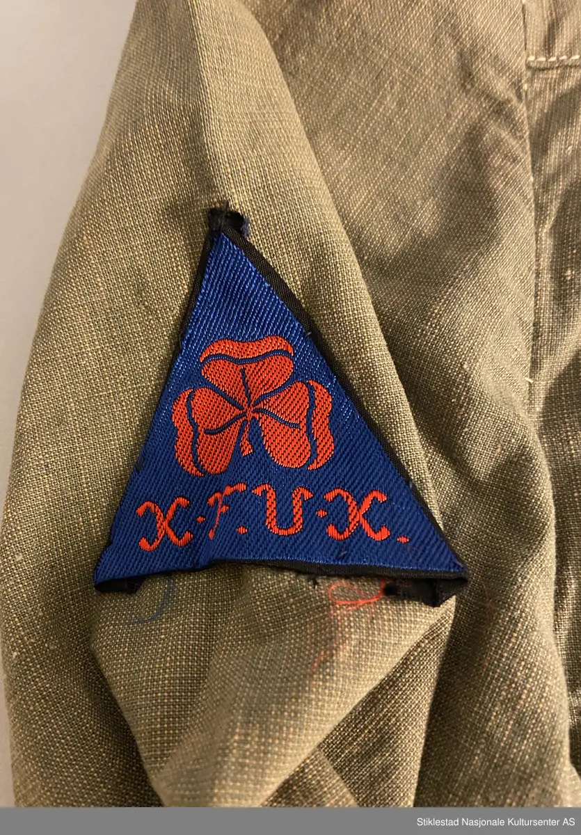 Speideruniform fra 1960-tallet. Dressen er i bomull og består av jakke, skjørt og lue (båtlue) i militærgrønn farge. Jakken har et påsydd stoffmerke på høyre arm: en blå trekant med trekløver og teksten KFUK - ("Kristelig Forening for Unge Kvinner"). På venstre skulderklaff er det festet et rødt bånd (dobbelt) med teksten «Verdal 1» (troppsgrad?). Rød snor i front for fløyte (denne mangler). Skjørtet er kort og "snurpet" sammen i midjen med strikk, to lommer foran. Merket K.K. på innsiden av ene lomme. Lue med påmontert merke i metall; trekløver i trekant. Innskripsjoner på innsiden av luen. Lue, militærgrønn, men annen grønn nyanse enn skjørt og jakke.