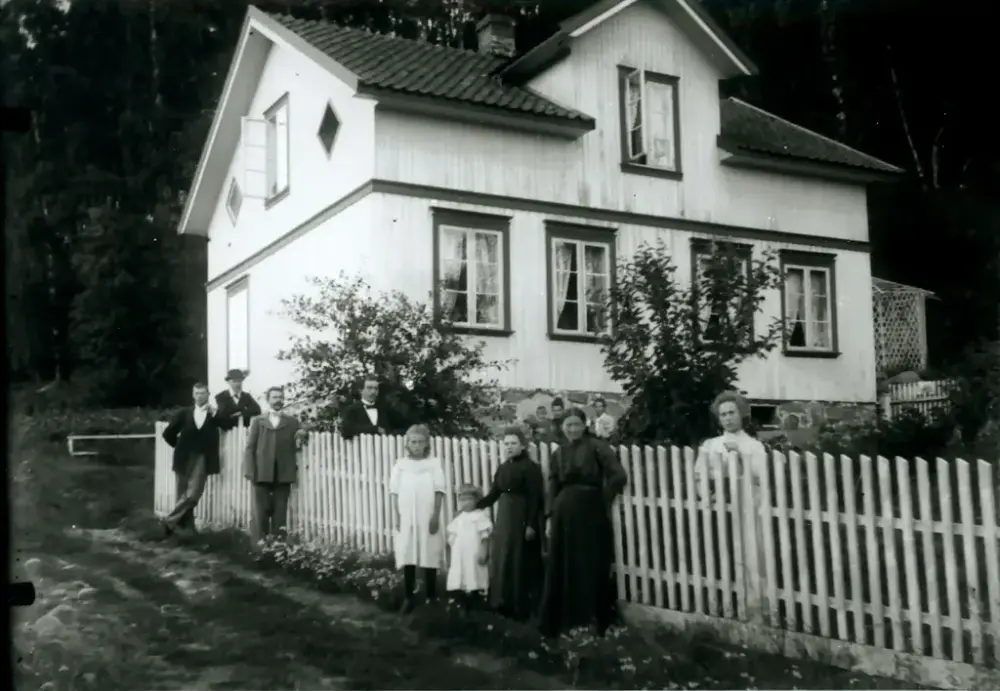 Et svart hvitt bilde av en sannsynlig familie på 12 foran et to etasjers hus med stakitt gjerdet