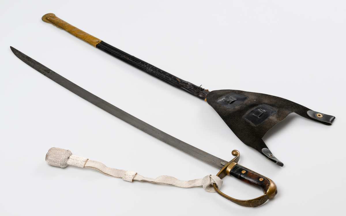 Polissabel m/26 med bäranordning. Handbygel och parérstång i förgylld mässing. Polisemblem på handbygeln. Kavel i läder. Handbygeln har en s.k. portepé i silverdrageri. Klinga i stål. Sabelbalja av läder, munbleck och beslag med släpsko i förgylld mässing.