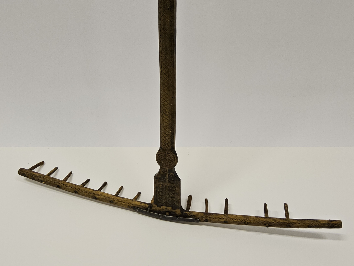 Räfsa med helt skaft. Rika sniderier, bl.a. rosett i karvsnitt samt stiliserade bladornament. På undersidan järnvägslatpp "Skultorp". Inköpt på auktion.