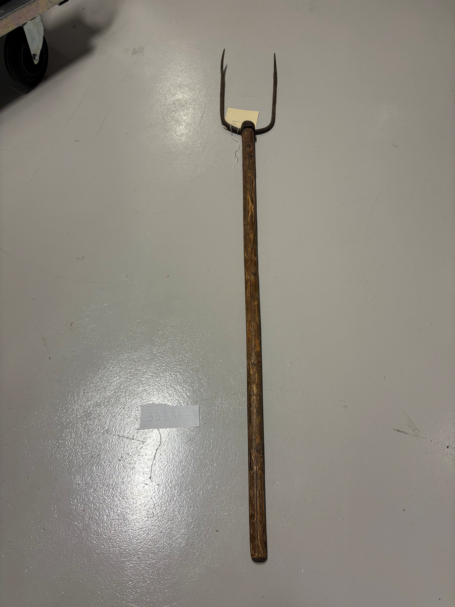 U-formad med två klor av järn fäst på skaft av trä med en järnten som utgår från gaffelns övre del. En järnring omsluter tenen och skaftet.