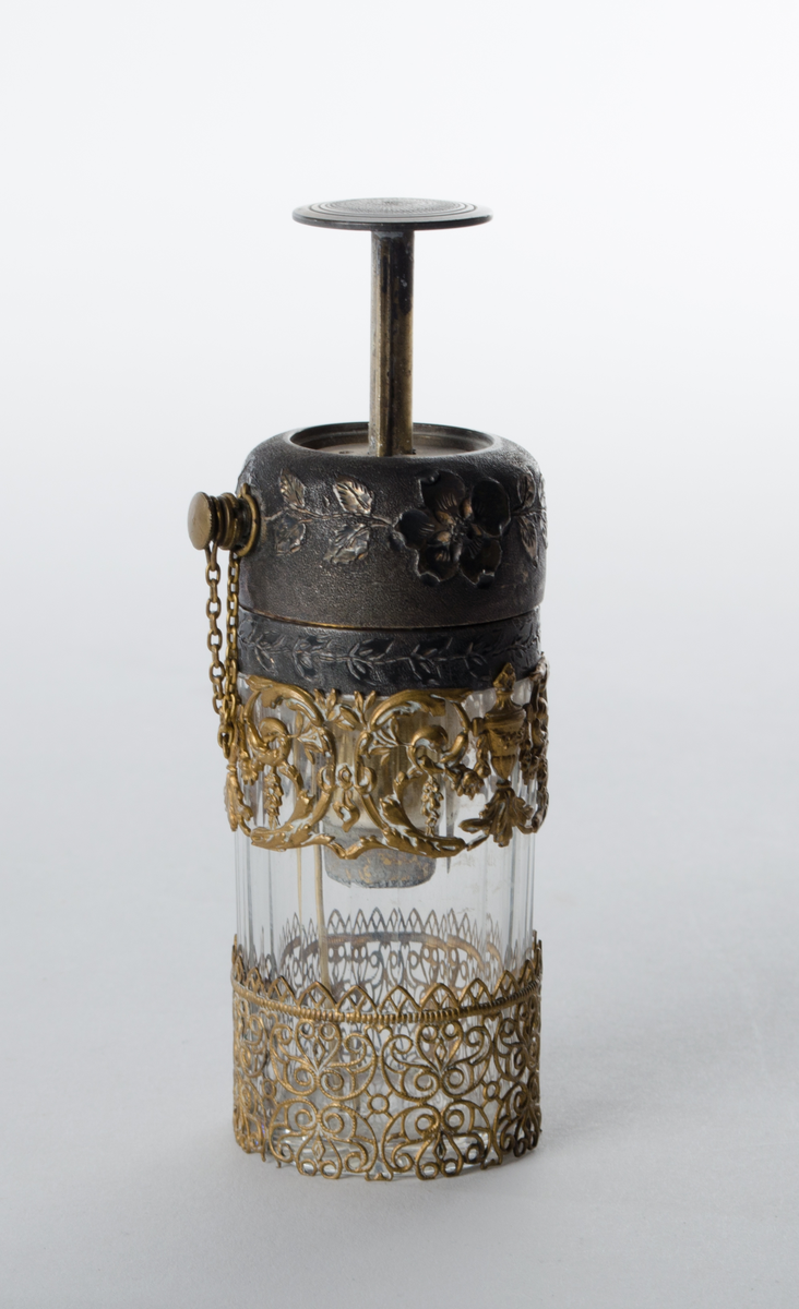 Pumpeflaske i glass med pumpemekanisme og spraytut i metall. Søvfarget og messingfarget dekor, fasettslipt glass. En av metallpyntringene er løse og har avvikende dekor (ikke original?)