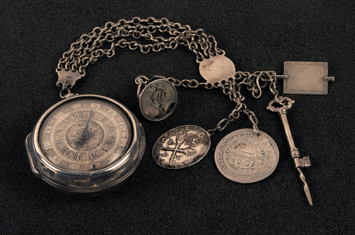 Lommeur med kjede av fire lenker og fem kjeder med anheng. Kjede med signet W, en oval medaljong med to kryssede fakler (WFA), en tannpirker formet som nøkkel, en mynt (F III 1666), firkantet uropptrekker. Stempel: se foto.