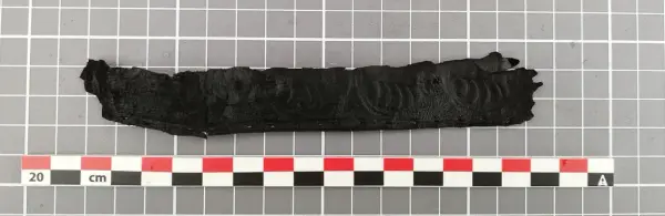 Avlangt fragment av mørkt lær med tydelig preget bølgelignende mønster, trolig fra en middelaldersk sverdslire.