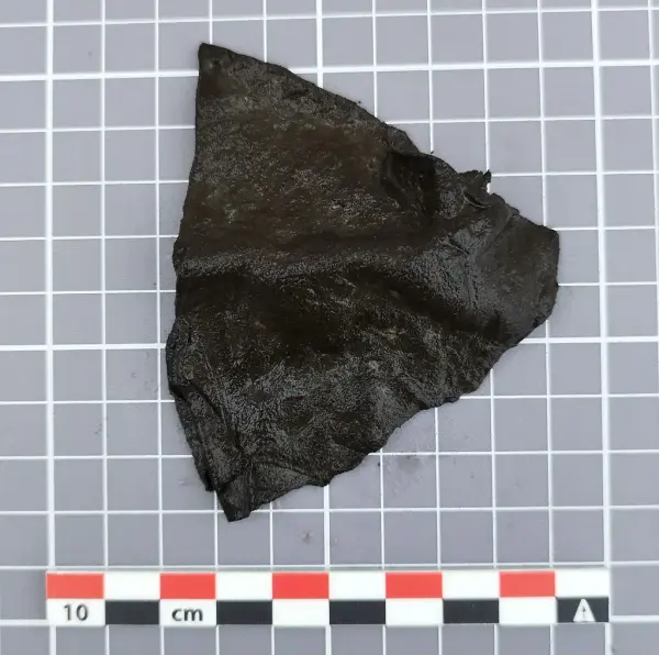 rekantet fragment av mørkt lær med bøyd fasong, trolig avskåret del av en middelaldersk lærslire.