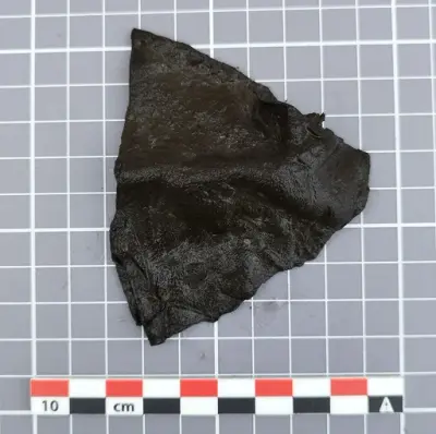 rekantet fragment av mørkt lær med bøyd fasong, trolig avskåret del av en middelaldersk lærslire.