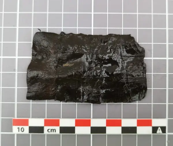  Lite, rektangulært fragment av mørkt lær med tydelige avkuttede kanter, trolig del av en middelaldersk slire.