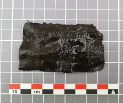  Lite, rektangulært fragment av mørkt lær med tydelige avkuttede kanter, trolig del av en middelaldersk slire.