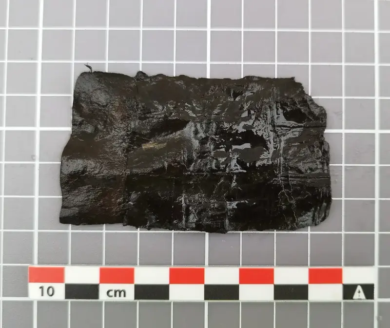  Lite, rektangulært fragment av mørkt lær med tydelige avkuttede kanter, trolig del av en middelaldersk slire.
