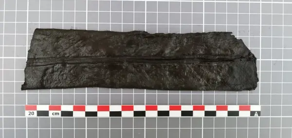 Mørkt lærfragment av en middelaldersk slire med tydelig gjennomgående søm midt på baksiden.