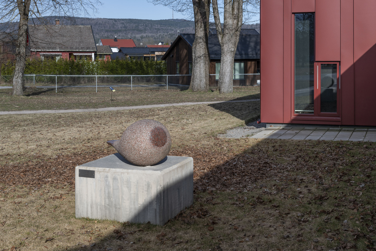 Skulptur utført i rød granitt. 140 cm lang med en tykk, rund del på ca 80 cm diameter og med en tynnere «hale». Et parti i den runde delen er polert slik at rødfargen er fremtredende.  