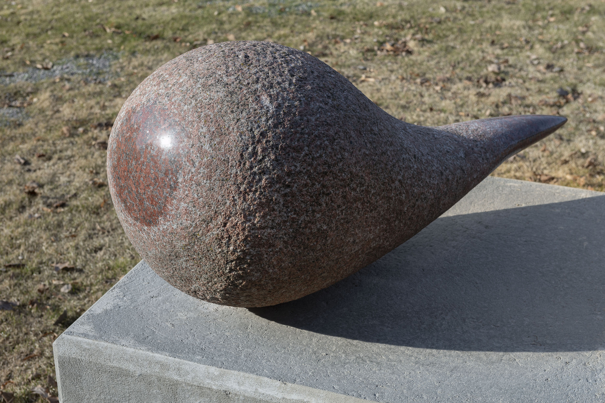 Skulptur utført i rød granitt. 140 cm lang med en tykk, rund del på ca 80 cm diameter og med en tynnere «hale». Et parti i den runde delen er polert slik at rødfargen er fremtredende.  