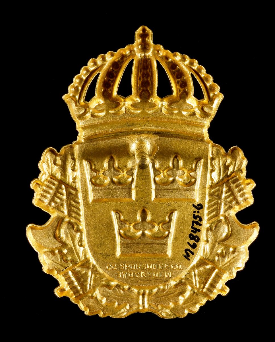 Mössplåt för polis. Sköld med lilla riksvapnet (tre kronor) med två bakomliggande fasces och ekkrans, krönt med kunglig krona. På frånsidan en fästskruv för montering.
