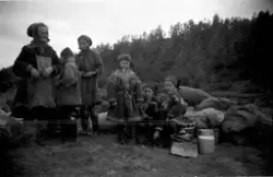 To samiske kvinner sammen med seks samiske barn i Kautokeino, 1955. Alle har kofta på seg. Sekker, kjeler, spann og reinskinn ligger på bakken. (Se også  bilde FBib.23013-119)